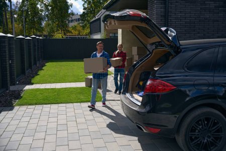 Photo for Full length photo of happy wife and husband carrying boxes to car with opened trunk while moving house - Royalty Free Image
