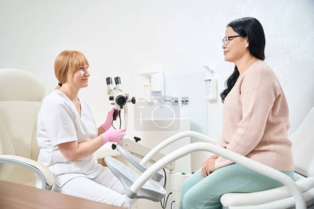 Ginecólogo experimentado hablando con paciente asiático adulto sobre su salud después del examen en silla ginecológica con microscopio, chequeo de salud de las mujeres