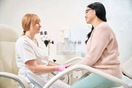 Woman gynecologist talking to her patient after examination on medical chair with microscope, specialist checking fallopian tubes patency and taking pap-test