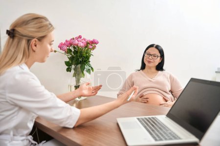 Foto de Mujer joven discutiendo los términos y el tamaño del bebé con la mujer adulta embarazada que entró en chequeo de salud de rutina, segundo trimestre, ginecología y reproductología - Imagen libre de derechos
