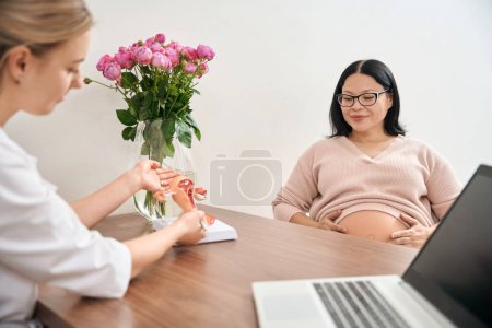 Téléchargez les photos : Qualified gynecologist showing to pregnant asian woman model of female vagina, explaining way of contractions and work of womb during baby delivering - en image libre de droit