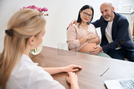 Téléchargez les photos : Couple de famille adulte visitant le bureau de l'obstétricien pour parler du troisième trimestre de la grossesse et du processus d'accouchement avec son aide, contracter l'accouchement - en image libre de droit