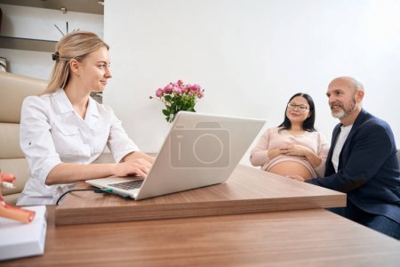 Photo for Young gynecologist filling in medical records of pregnant woman on laptop, listening to client complains and feelings, checking baby well-being, routine health check-up - Royalty Free Image