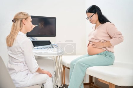 Photo for Pregnant asian woman visiting sonographer for extra ultrasound, doctor checking health of unborn baby and pointing at image in the womb on digital display - Royalty Free Image