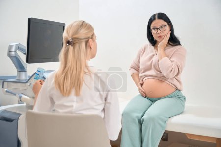 Surprised pregnant woman consulting with gynecologist-obstetrician about ways of childbirth after ultrasonography, contemplating a caesarean section birth