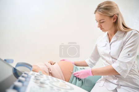 Téléchargez les photos : Jeune femme médecin faisant l'injection à la femme enceinte qui se trouve sur le canapé médical, injections d'immunoglobuline Rh en raison du risque d'incompatibilité Rh - en image libre de droit