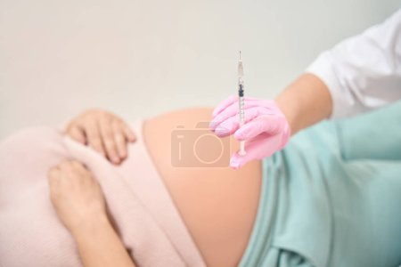 Photo for Person in white robe and safety gloves holding syringe with liquid, pregnant woman lying on the background, Rh immunoglobulin injection, gynecology and reproductology - Royalty Free Image