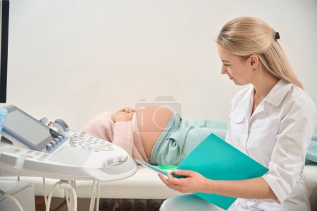 Photo for Smiling woman ob-gyn writing down medical records after ultrasound of pregnant woman, inserting data about fetus size and presentation inside the woman womb - Royalty Free Image
