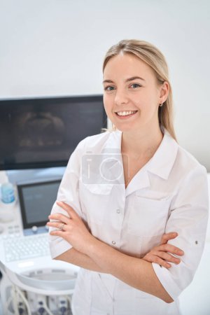 Pretty high qualified woman sonographer smiling standing near the ultrasound machine, clinic suggesting all types of ultrasounds for people, healthcare and medicine