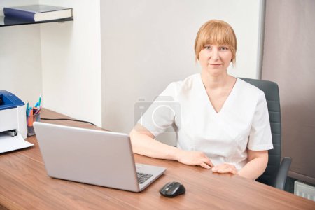 Femme médecin hautement qualifiée attendant un client assis dans un lieu de travail confortable avec ordinateur portable, effectuant des examens médicaux, travaillant dans une clinique privée, les soins de santé et la médecine