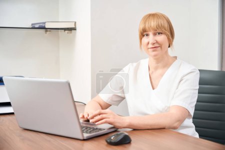 Smiling woman therapist keeping online medical consultation to her patient, checking ill patients and prescribing additional medicine using laptop in her office