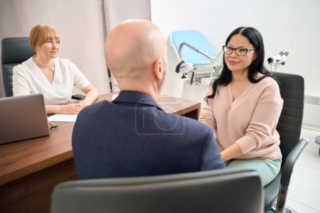 Happy asian woman looking at her husband with love and gratefulness sitting in gynecologist office, man agreeing to get a baby in the way of artificial fertilization