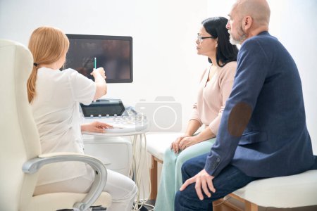 Foto de Médico altamente calificado que habla con los futuros padres después de la ecografía, señala al feto en la pantalla digital y explica los riesgos del embarazo en la edad adulta - Imagen libre de derechos