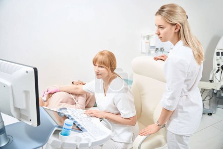 Woman sonographer performing ultrasound of the thyroid glands to woman who going to get pregnant, female assistant helping her to analyze data, health check-up