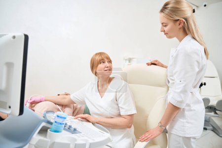 Diplom-Sonografin erklärt der jungen Krankenschwester, wie man den Schilddrüsen-Ultraschall macht, Patientin liegt auf medizinischem Sofa in Arztpraxis