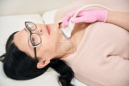 Téléchargez les photos : Gros plan femme asiatique dans les lunettes couché sur le canapé de la procédure médicale tandis que le médecin échographe effectuer une échographie de sa glande thyroïde - en image libre de droit