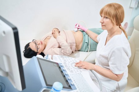 Photo for Serious woman gynecologist examining pregnant woman, checking the position and health condition of baby in patient womb, making ultrasound in private clinic - Royalty Free Image