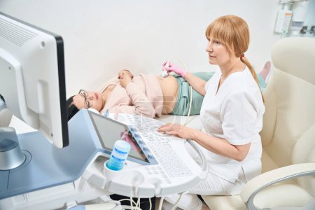 Photo for High-qualified gynecologist performing extra fetal ultrasound to adult pregnant woman because of such her conditions like age over 40 and extra weight, health check-up - Royalty Free Image