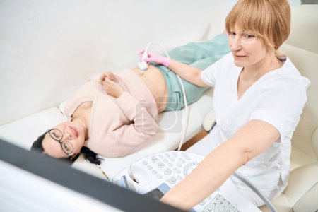 Erwachsene asiatische Frau bekommt Ersttrimester-Ultraschall, um das Vorhandensein einer Schwangerschaft zu überprüfen, Sonografin zeigt den Fötus auf der digitalen Anzeige des Ultraschallgeräts