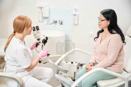 Female gynecologist preparing gynecological microscope to check female patient who sitting on gynecological examination chair health of reproductive organs, pelvic exam
