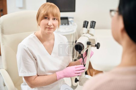 Ginecólogo médico calificado va a hacer examen ginecológico de paciente mujer en silla con microscopio ginecológico, chequeo de salud de las mujeres