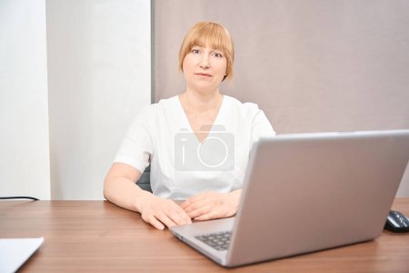 Téléchargez les photos : Médecin généraliste assis sur le bureau avec ordinateur portable dans son bureau, attendant le client, menant des consultations médicales en ligne, les soins de santé et la médecine - en image libre de droit