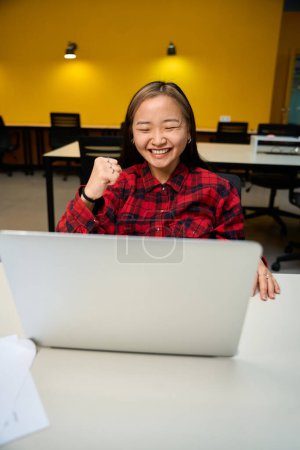 Téléchargez les photos : Jeune asiatique heureuse employée de l'informatique célébrant le succès et l'utilisation de l'ordinateur portable au bureau dans le bureau de coworking. Concept de travail indépendant ou à distance moderne - en image libre de droit