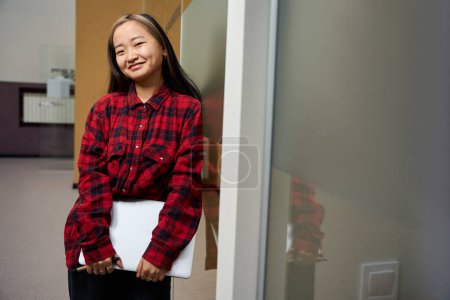 Téléchargez les photos : Jeune asiatique souriante employée informatique avec ordinateur portable regardant caméra par mur de verre dans le bureau de coworking. Concept de travail indépendant ou à distance moderne - en image libre de droit
