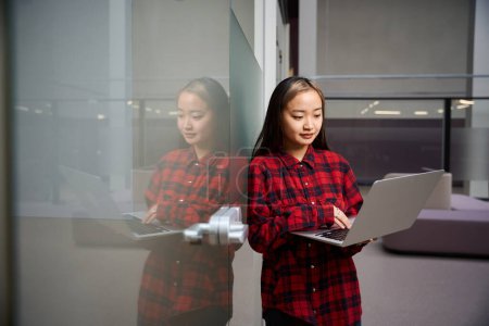 Téléchargez les photos : Jeune asiatique ciblée employée informatique utilisant ordinateur portable par mur de verre dans le bureau de coworking. Concept de travail indépendant ou à distance moderne - en image libre de droit