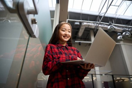Téléchargez les photos : Vue du bas de jeune asiatique souriante employée des TI regardant ordinateur portable par mur de verre dans le bureau de coworking. Concept de travail indépendant ou à distance moderne - en image libre de droit