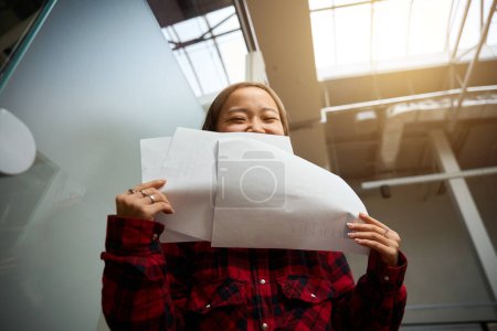 Téléchargez les photos : Vue du bas de la jeune employée asiatique en informatique couvrant la moitié du visage avec des papiers dans un bureau de coworking flou. Concept de travail indépendant ou à distance moderne - en image libre de droit