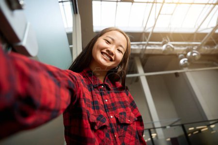 Téléchargez les photos : Vue en angle élevé de la jeune employée asiatique souriante en informatique regardant la caméra dans un bureau de coworking flou. Concept de travail indépendant ou à distance moderne - en image libre de droit