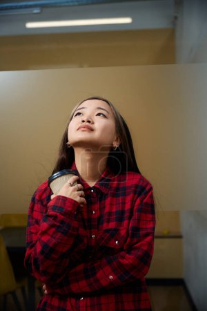 Photo for Young thoughtful asian female IT employee with coffee cup by glass wall in coworking office. Concept of modern freelance or remote work - Royalty Free Image