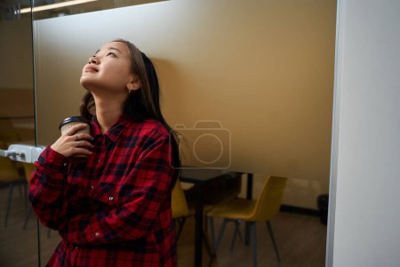 Téléchargez les photos : Jeune asiatique travailleuse informatique avec tasse de café penser à quelque chose par mur de verre dans le bureau de coworking. Concept de travail indépendant ou à distance moderne - en image libre de droit