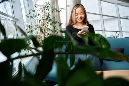 Foto de Joven sonriente asiática empleada de TI utilizando smartphone en sofá en la oficina de coworking. Concepto de trabajo independiente o remoto moderno - Imagen libre de derechos