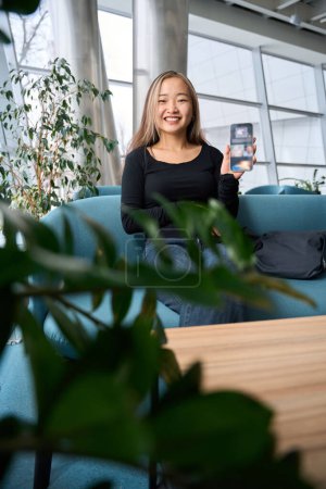 Téléchargez les photos : Jeune asiatique souriante employée informatique montrant quelque chose sur smartphone à la caméra sur canapé dans le bureau de coworking. Concept de travail indépendant ou à distance moderne - en image libre de droit