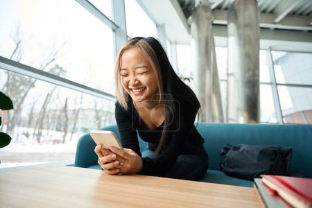 Photo for Young laughing asian female IT employee using smartphone on sofa at table in coworking office. Concept of modern freelance or remote work - Royalty Free Image
