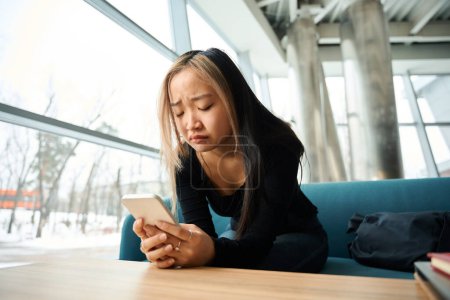 Téléchargez les photos : Jeune femme asiatique triste employé de l'informatique en utilisant un smartphone sur le canapé à la table dans le bureau de coworking. Concept de travail indépendant ou à distance moderne - en image libre de droit