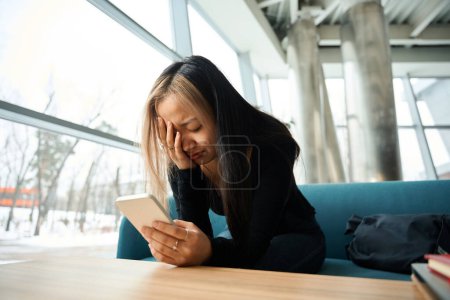 Téléchargez les photos : Jeune asiatique bouleversée et pleurant employée de l'informatique en utilisant un smartphone sur le canapé à la table dans le bureau de coworking. Concept de travail indépendant ou à distance moderne - en image libre de droit