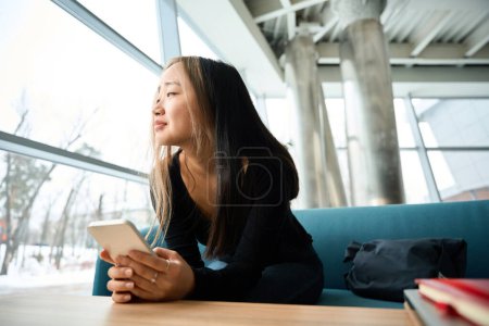 Photo for Young sad asian female IT employee with smartphone looking away on sofa at table in coworking office. Concept of modern freelance or remote work - Royalty Free Image