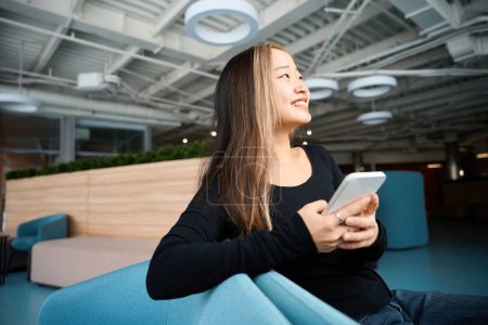 Foto de Joven sonriente asiática gerente de TI femenina con teléfono inteligente mirando hacia otro lado en el sofá en la oficina de coworking. Concepto de trabajo independiente o remoto moderno - Imagen libre de derechos