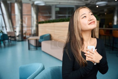 Photo for Young thoughtful asian female IT employee holding smartphone on sofa in blurred coworking office. Concept of modern freelance or remote work - Royalty Free Image