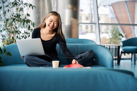 Foto de Joven riendo asiática empleada de TI femenina tomando cuaderno mientras trabaja en el ordenador portátil en el sofá en la oficina de coworking. Concepto de trabajo independiente o remoto moderno - Imagen libre de derechos