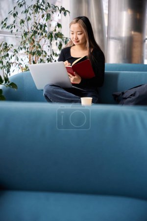 Photo for Young focused asian female IT employee writing in notebook and watching laptop on sofa in coworking office. Concept of modern freelance or remote work - Royalty Free Image