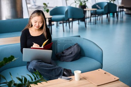 Foto de Joven asiática concentrada gerente de TI escribiendo en el cuaderno y viendo portátil en el sofá en la oficina de coworking. Concepto de trabajo independiente o remoto moderno - Imagen libre de derechos