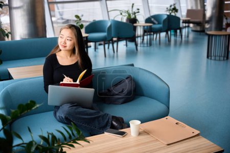 Photo for Young thoughtful asian female IT employee looking away while working on laptop and writing in copybook on sofa in coworking office. Concept of modern freelance or remote work - Royalty Free Image