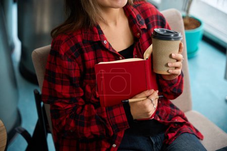 Photo for Focus on foreground of partial female IT employee with coffee cup and notebook on chair in blurred coworking office. Concept of modern freelance or remote work - Royalty Free Image