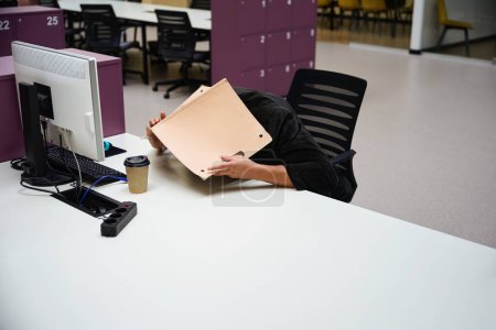 Photo for Copy space photo of male employee sitting in coworking space while lying on table and covering head with folder - Royalty Free Image
