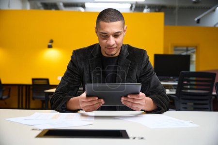 Photo for Smiling young man sitting at table in call center and holding tablet in hands while having online conversation - Royalty Free Image