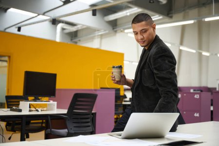 Photo for Copy space photo of smiling young male employee standing in call center while drinking coffee and watching something on laptop - Royalty Free Image